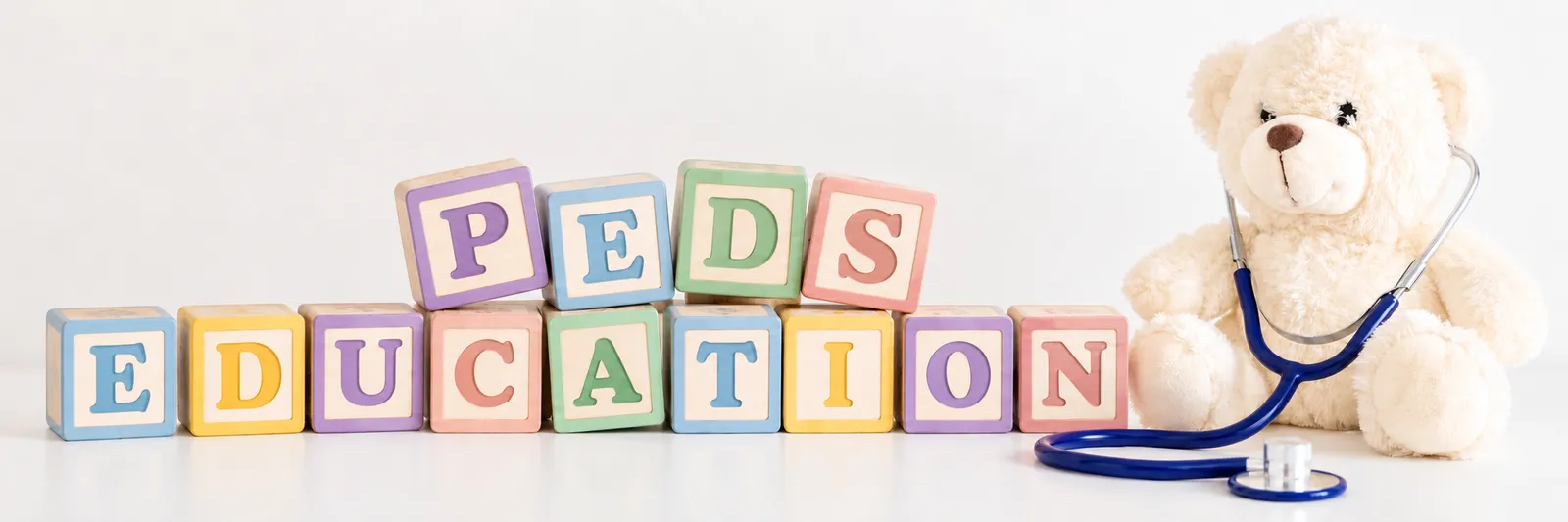 Wooden alphabet blocks spelling PEDS EDUCATION beside a teddy bear wearing a stethoscope